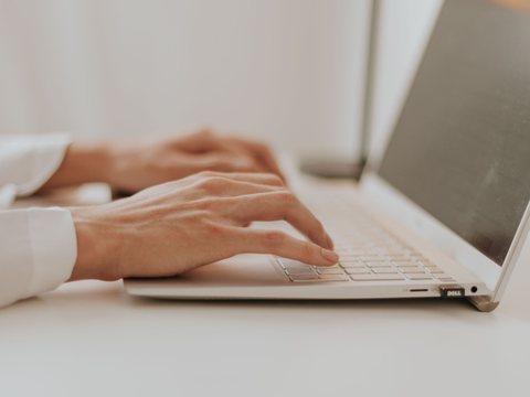 Mãos digitando em um notebook, perto de uma janela iluminada